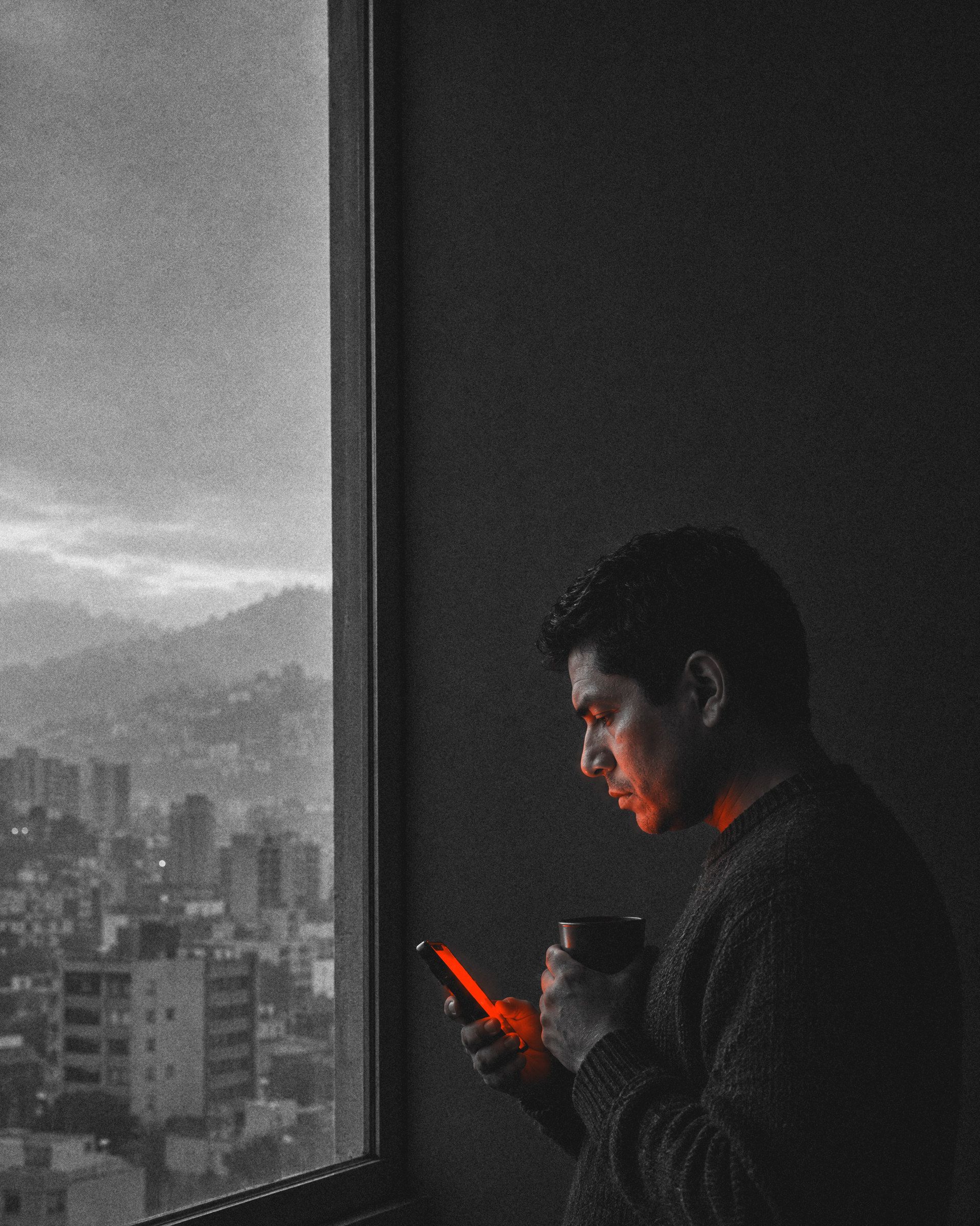 Homem latino-americano de suéter escuro, de perfil junto à janela ao amanhecer, segura um café e lê o celular. Metade do rosto está na sombra; a tela ilumina a mão e o rosto de vermelho. Lá fora, prédios e morros na névoa.